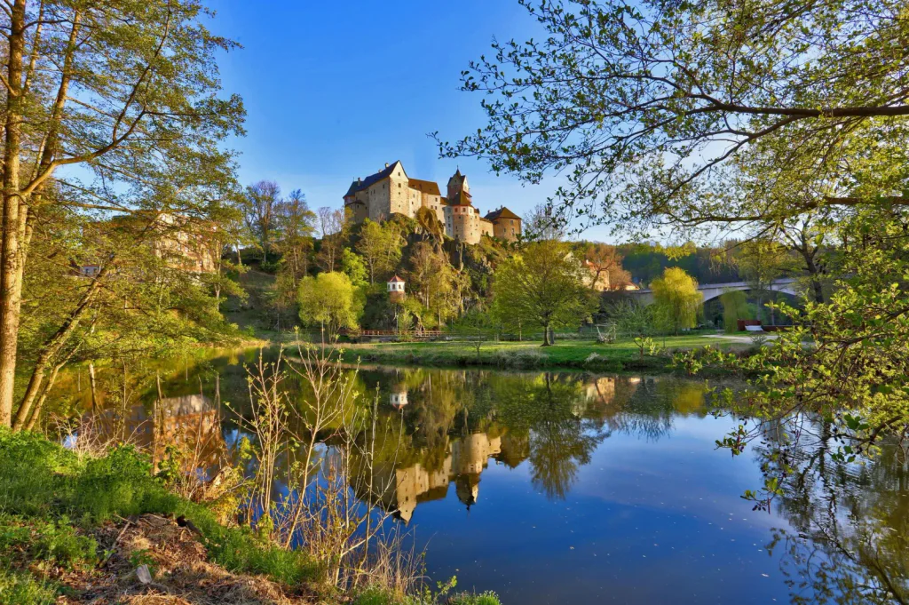 Pohled zpoza jezera na hrad Loket zasazený v malebné krajině.