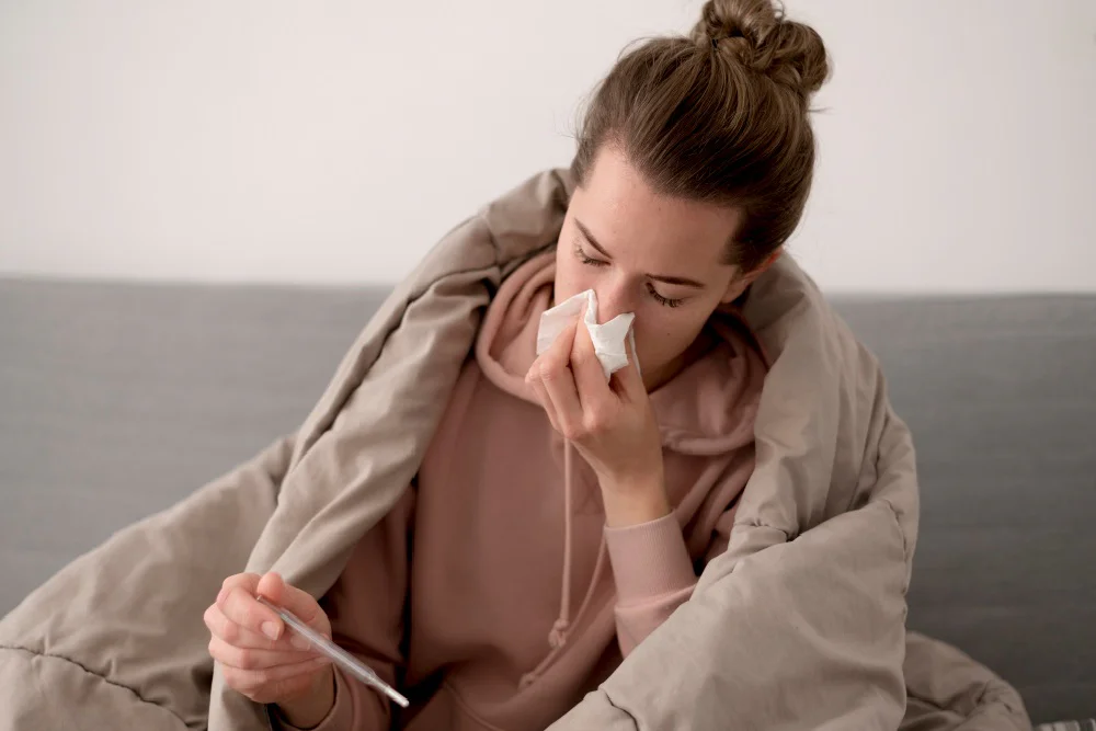 Smrkající žena s dekou na zádech se dívá na teploměr