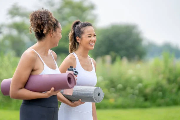 Two women in athletic wear walk outdoors with rolled yoga mats, smiling and chatting in a park setting.