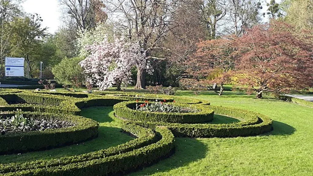 Podzámecké zahrada v Kroměříži je upravena do kulatých a elipsovitých tvarů v kterých se nacházejí květiny. V pozadí se nachází zelené stromy, z nichž jeden je bílý a druhý červený v krásném slunečném dni
