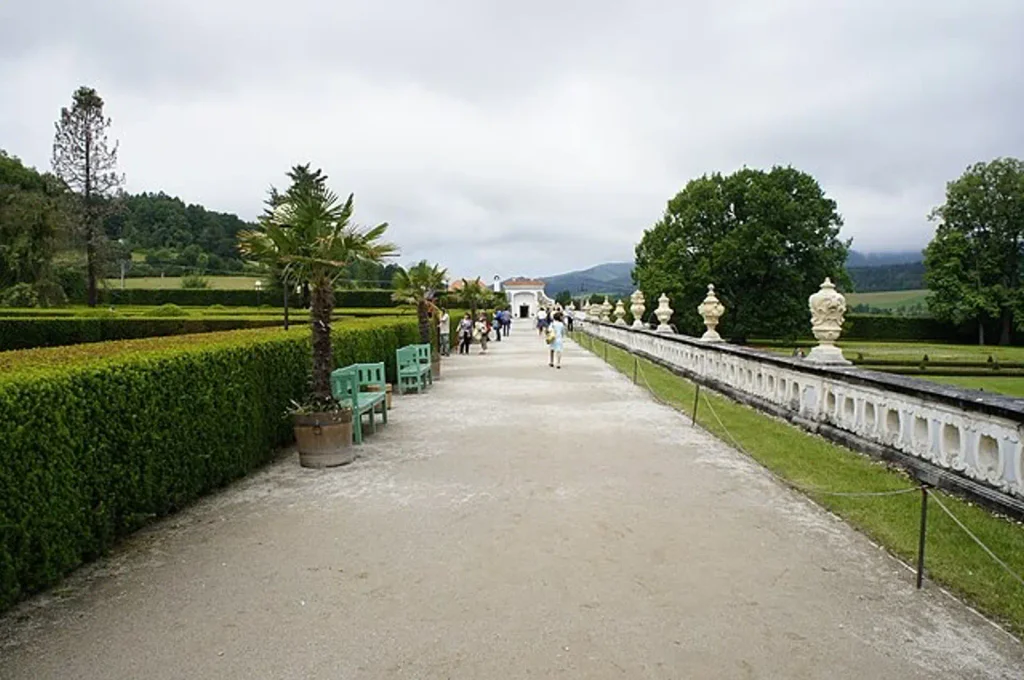 Dlouhý chodník v zámecké zahradě v Českém Krumlově, na levé straně se nachází zelený živý plot s dvěma zeleným lavičkami. Na pravé straně je bílo-šedý betonový zdobený plot. V pozadí jsou stromy s polojasnou oblohou