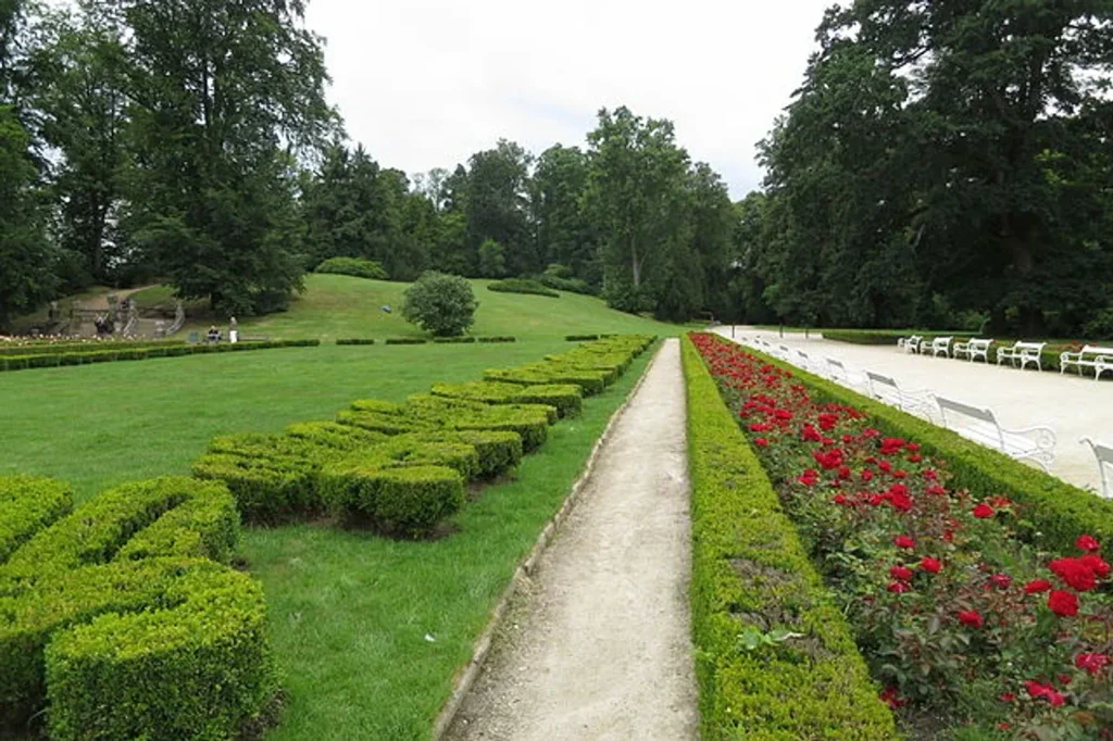 Uprostřed zámeckého parku v Hluboké pod Vltavou vede úzký chodník, obklopený na levé straně různými tvary vystříhaných z nízkých keřů. Na pravé straně se nachází dlouhý záhon červených květin. Pozadí obklopuje kopec a zelené stromy 