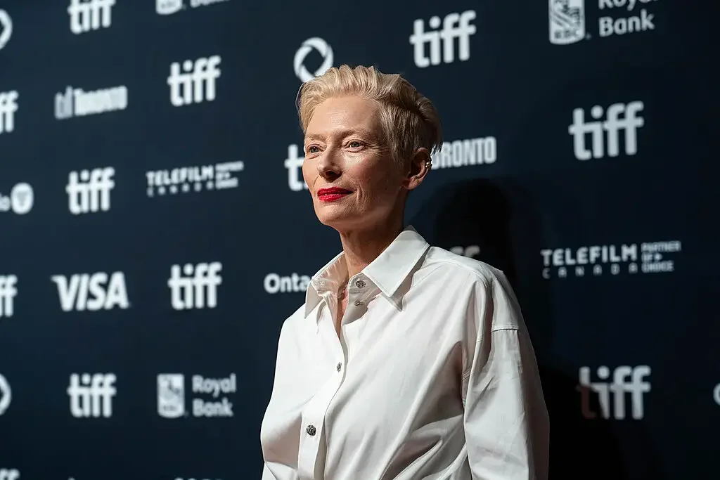 Tilda Swinton v bílé košili a s červenou rtěnkou pózuje před stěnou filmového festivalu