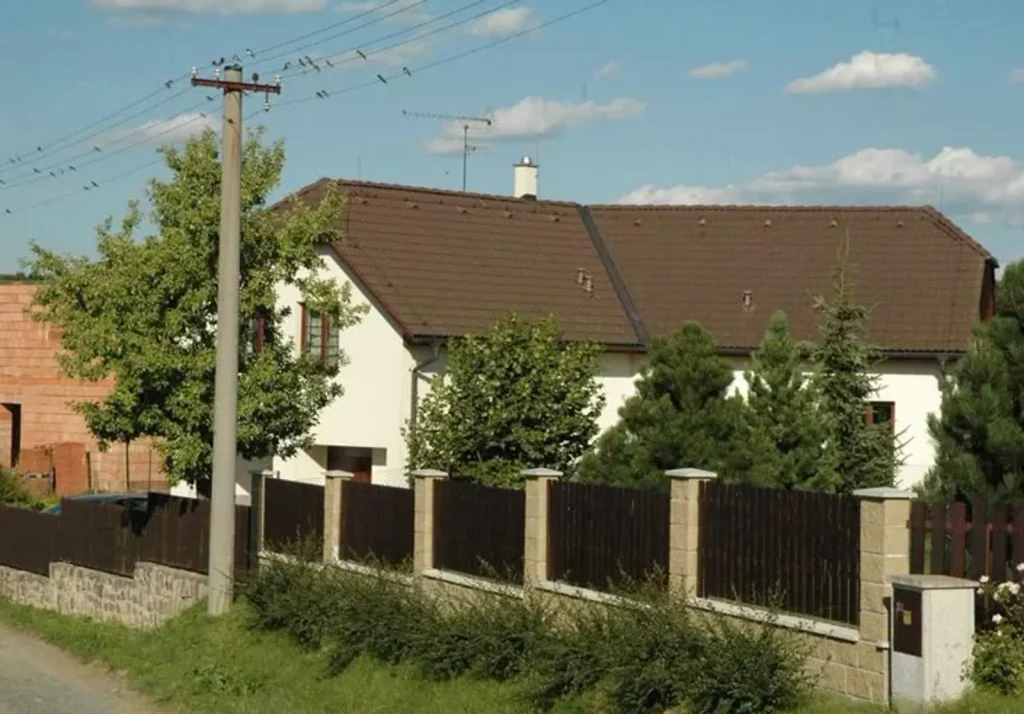 Hercova bílá chalupa s hnědou střechou, která je schována za dřevěný plot a část jehličnatých stromů