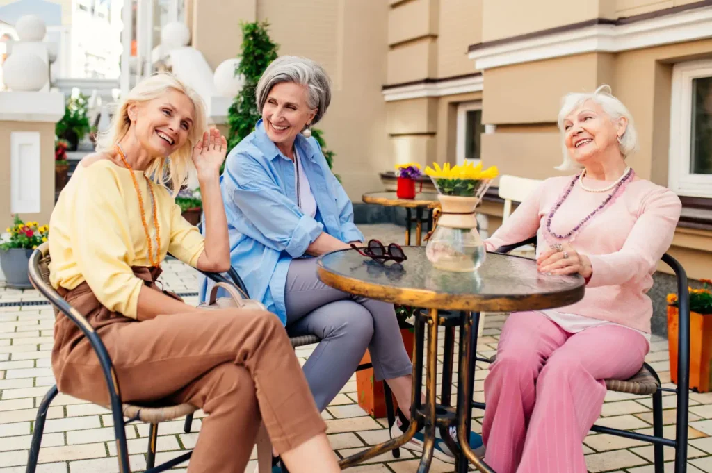 Tři elegantně oblečené ženy středního a vyššího věku u kavárenského stolku, smějí se a příjemně spolu tráví čas venku.