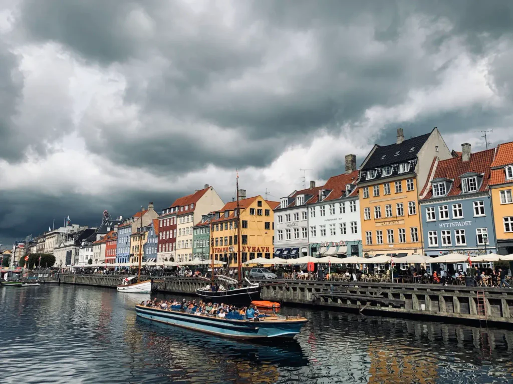 Záběr na hlavní město Dánska, Kodaň. Vidět je kanál s vodou, na kterém pluje loď a na břehu ho lemují barevné domky.