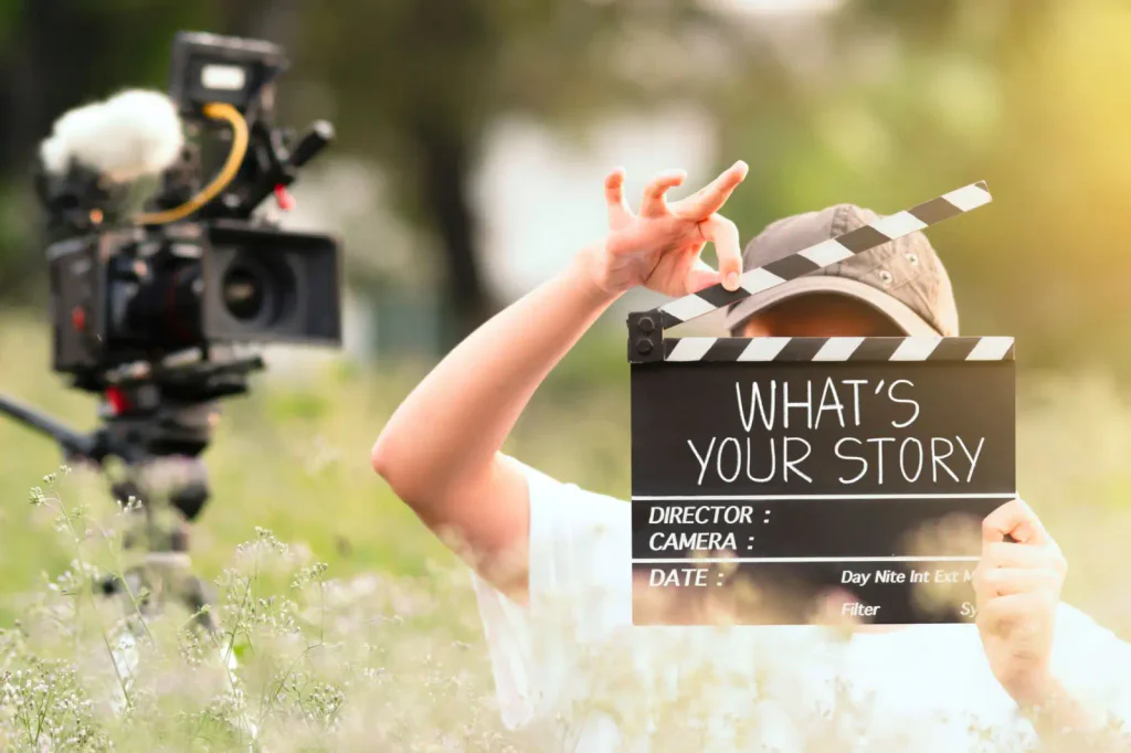 Fotka filmové klapky, kterou drží dítě. Nalevo vedle toho je profesionální kamer na stativu.